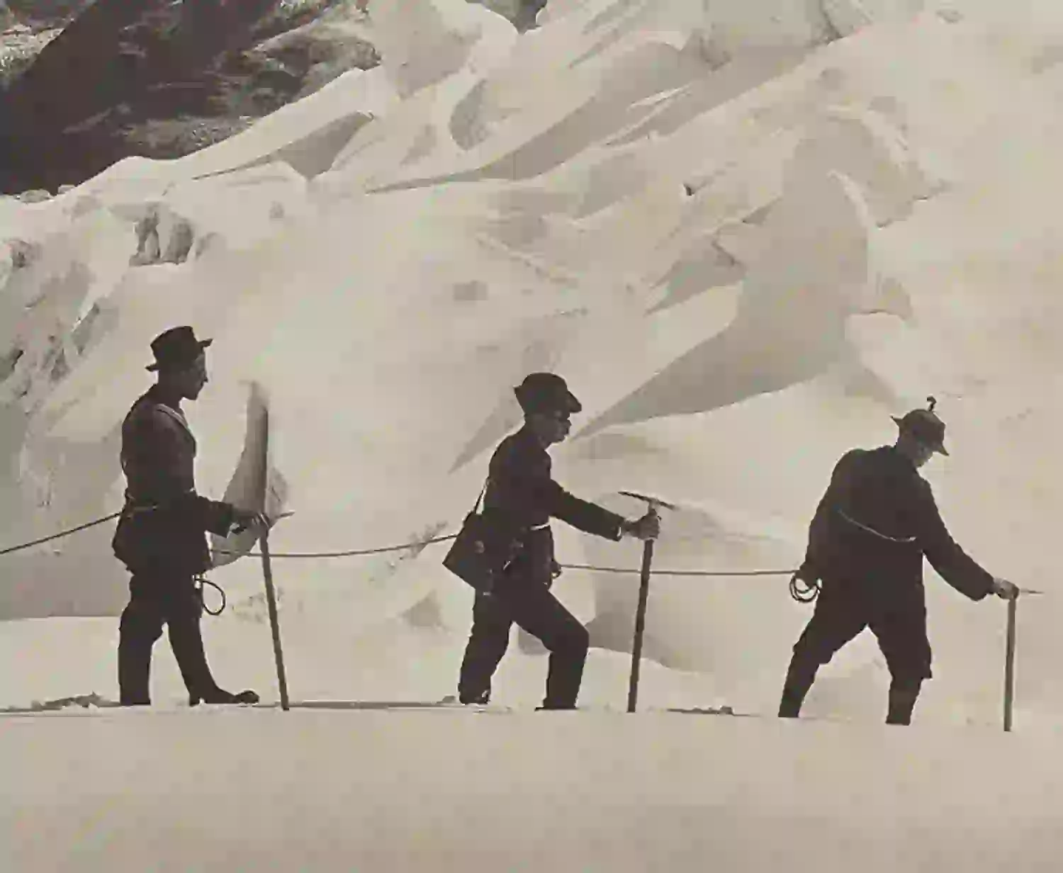 Drei aneinander geseilte Bergsteiger mit Hüten wandern im Gänsemarsch durch eine verschneite, eisige Landschaft mit schroffen, sonnenbeschienenen Gletscherformationen im Hintergrund.