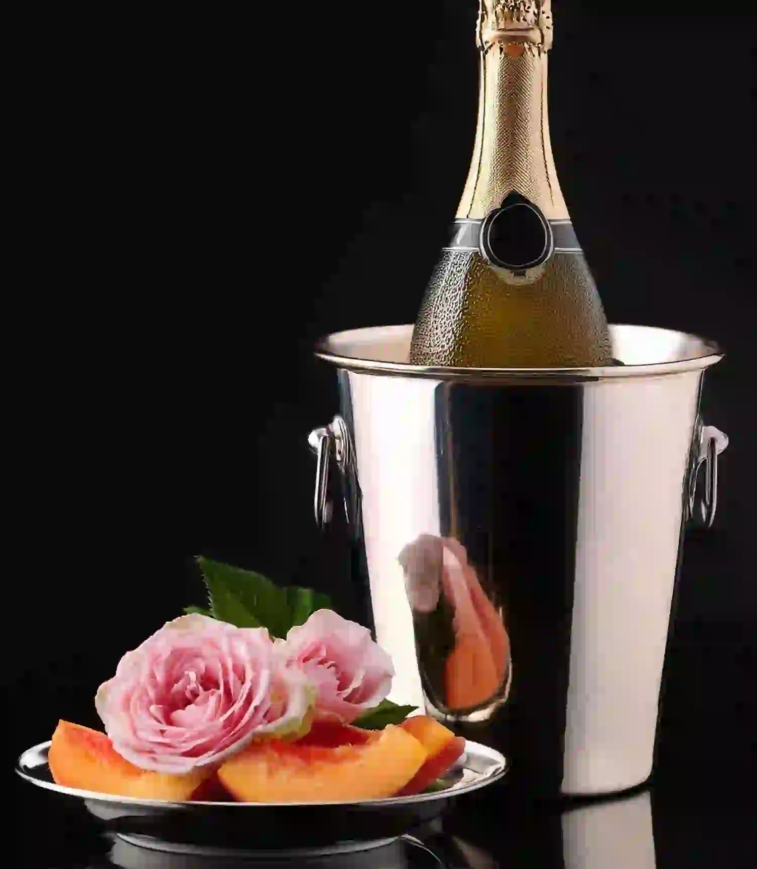 Eine Flasche Champagner in einem silbernen Eiskübel steht hinter einem Teller mit Pfirsichscheiben und rosa Rosen, alles vor einem schwarzen Hintergrund – perfekt für luxuriöse Verwöhnpakete.