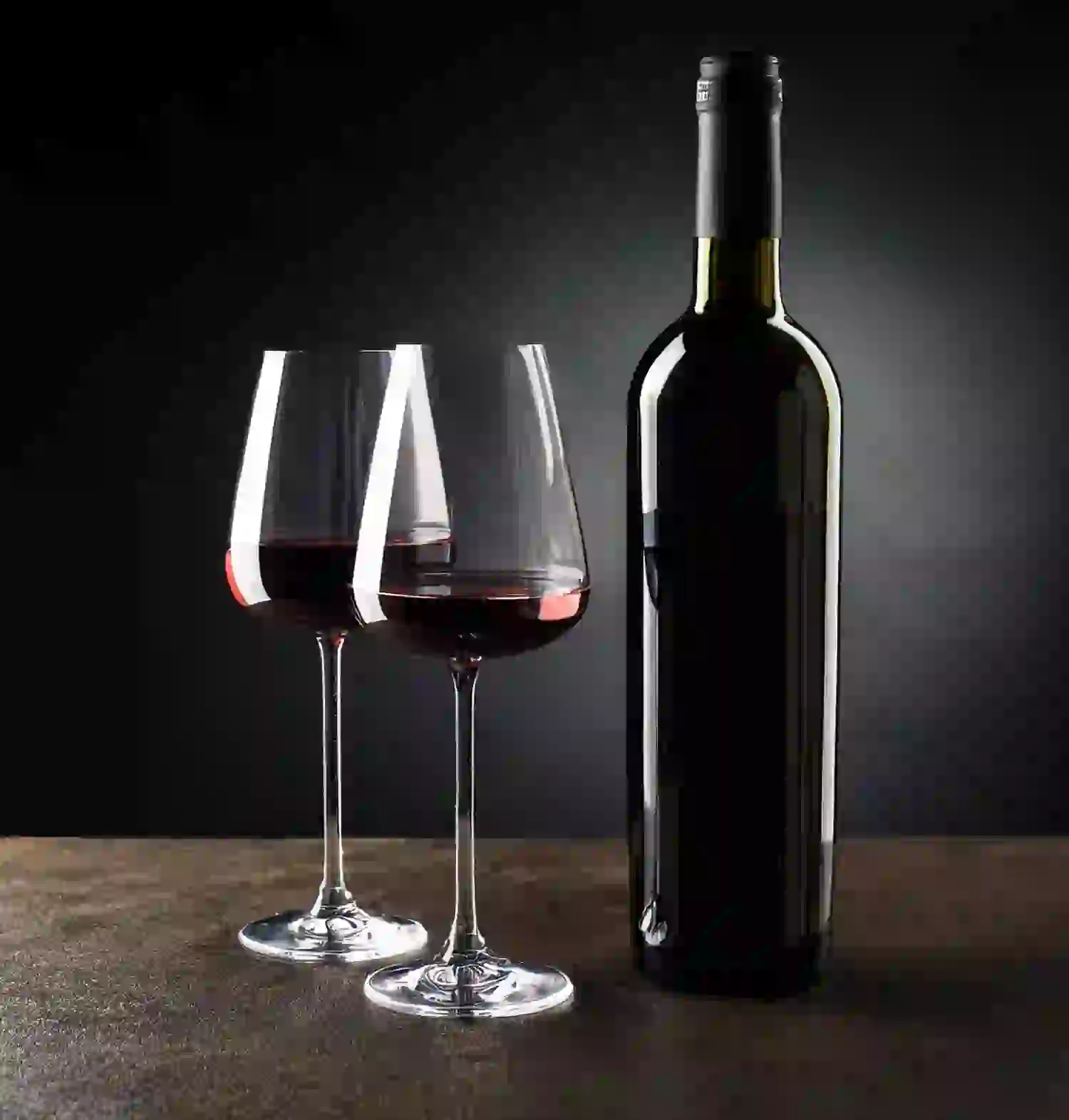 Eine dunkle Weinflasche mit schwarzem Verschluss steht neben zwei hohen, mit Rotwein gefüllten Gläsern auf einer dunklen Oberfläche und erinnert vor einem schwarzen Hintergrund mit Farbverlauf an die Eleganz exklusiver Verwöhnpakete.