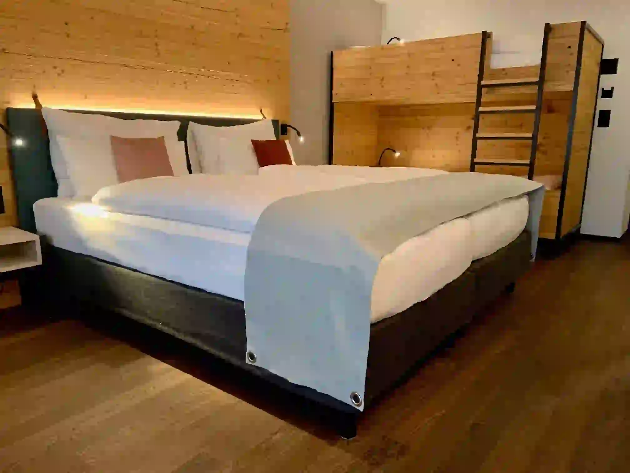 A cozy hotel room with a large double bed dressed in white linens and a light gray runner, next to wooden bunk beds. The room features wood-paneled walls and flooring, creating a warm atmosphere.