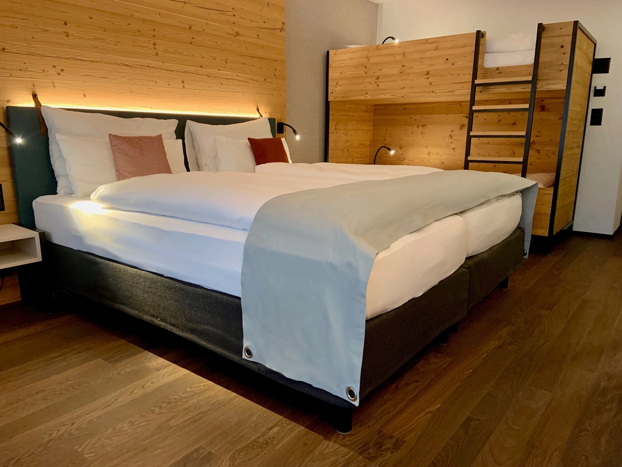 Ein gemütliches Hotelzimmer mit einem großen, hübsch gemachten Doppelbett mit weißer Bettwäsche und grauer Decke, sanfter Beleuchtung, Holzwänden, dunklem Holzboden und einem Etagenbett aus Holz mit einer Leiter in der Ecke.