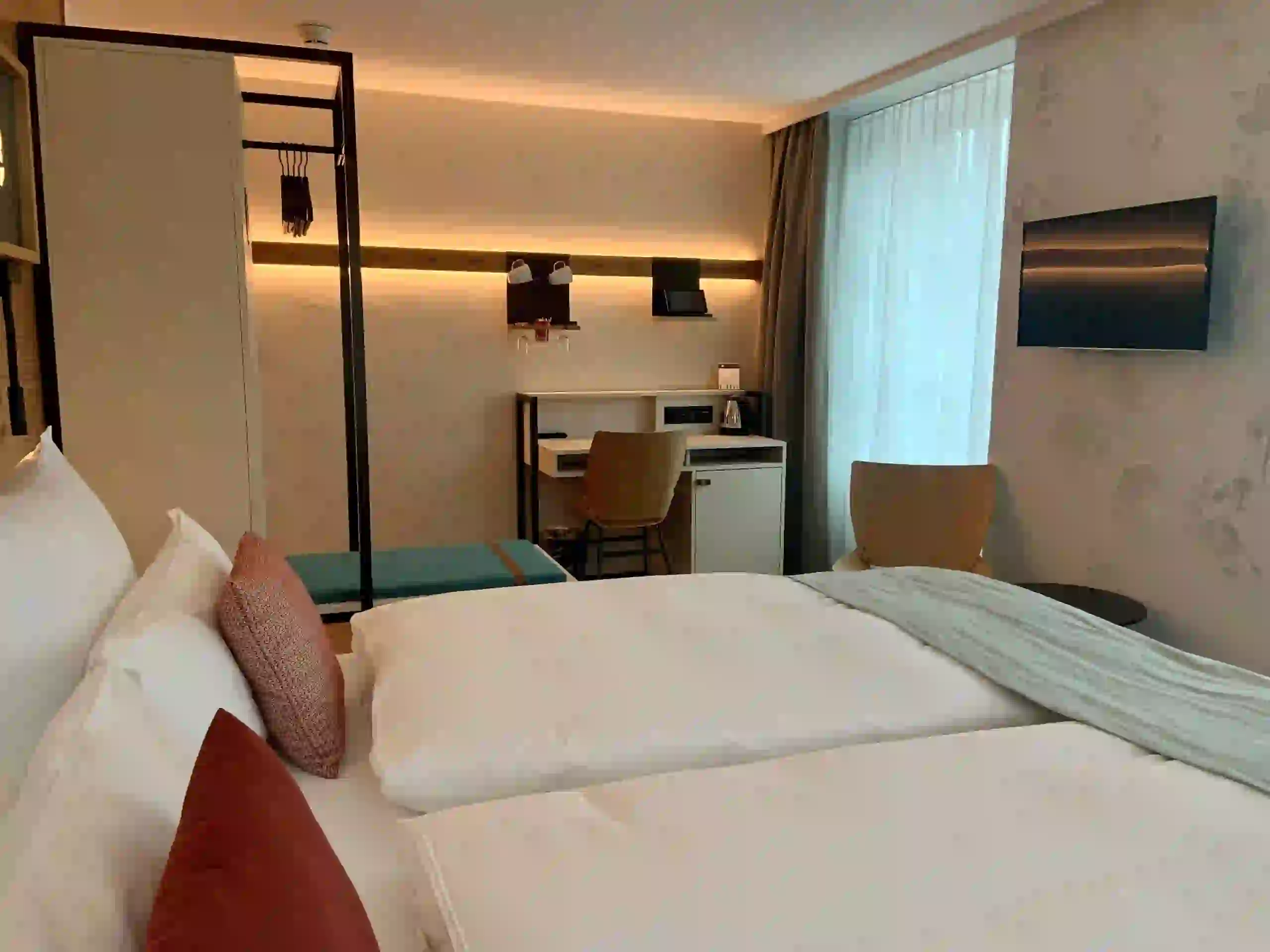 A modern hotel room with neatly made twin beds, a small desk and chair, a wall-mounted TV, an open wardrobe, and a window with sheer curtains letting in natural light.