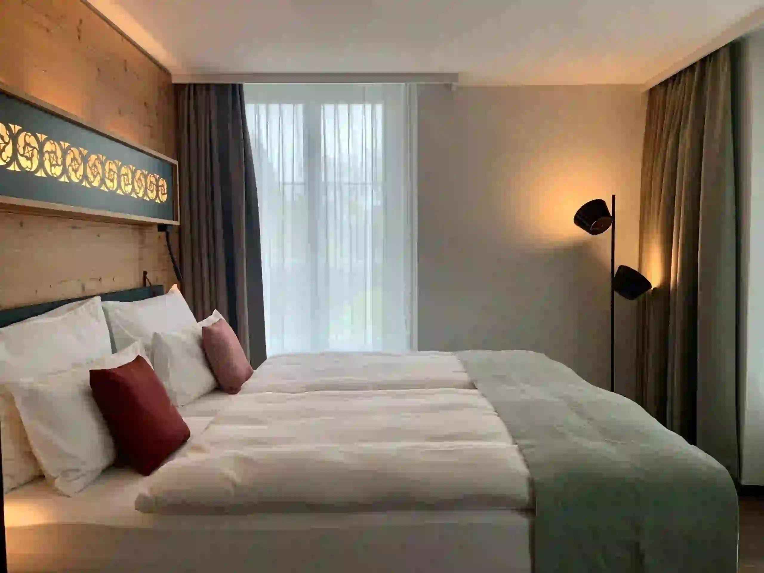 Modern hotel room with a large neatly made bed, white linens, red accent pillows, and a green throw. Soft lighting from a standing lamp, wooden and neutral walls, and sheer curtains covering a large window create a cozy atmosphere.