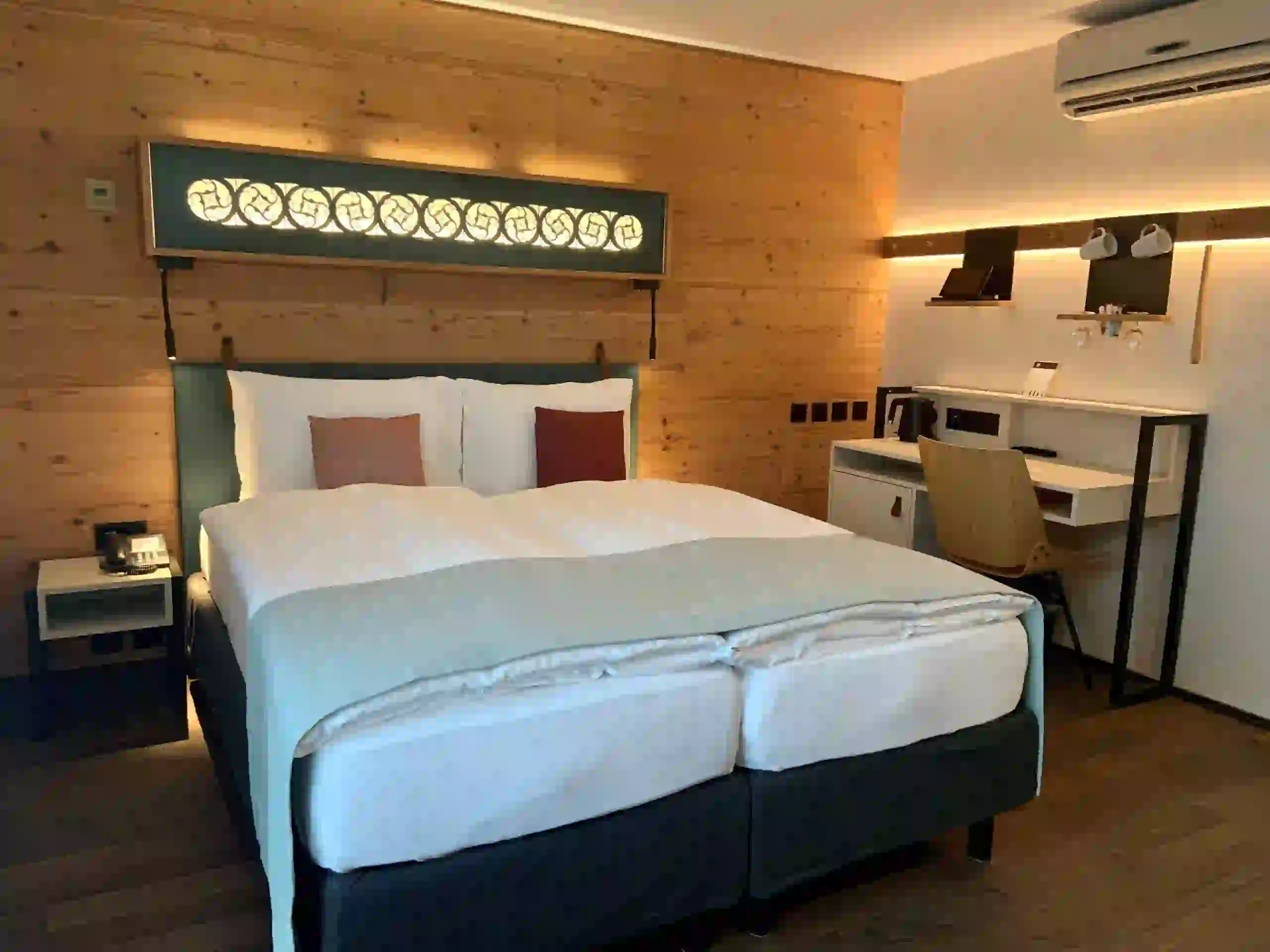 A modern hotel room with a double bed, wooden walls, soft lighting, a desk with a chair, a wall-mounted air conditioner, and neatly arranged amenities.