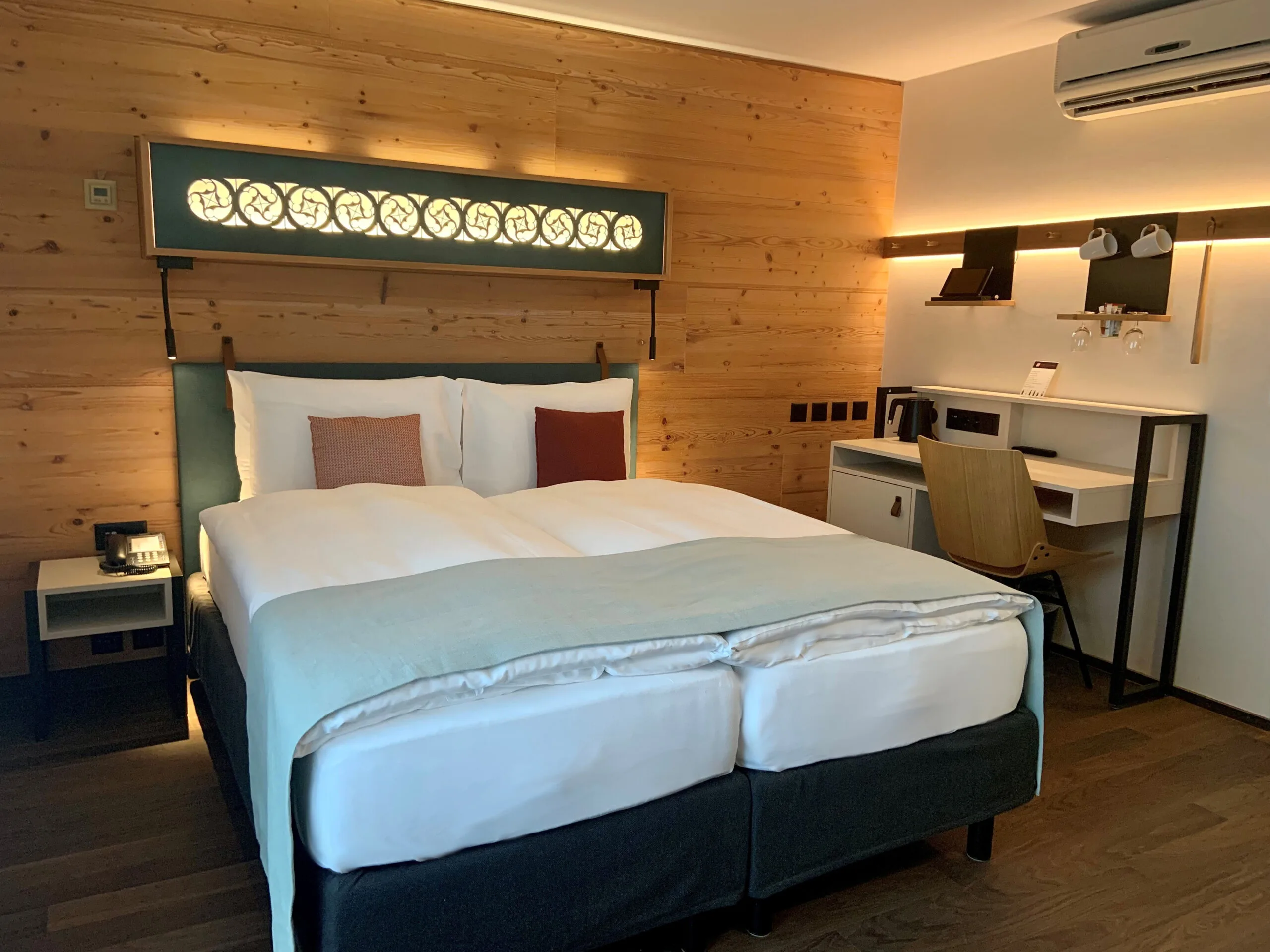 Ein gemütliches Hotelzimmer mit einem Doppelbett, holzgetäfelten Wänden, einem modernen Schreibtisch mit Stuhl, einer an der Wand montierten Klimaanlage und warmer Umgebungsbeleuchtung.