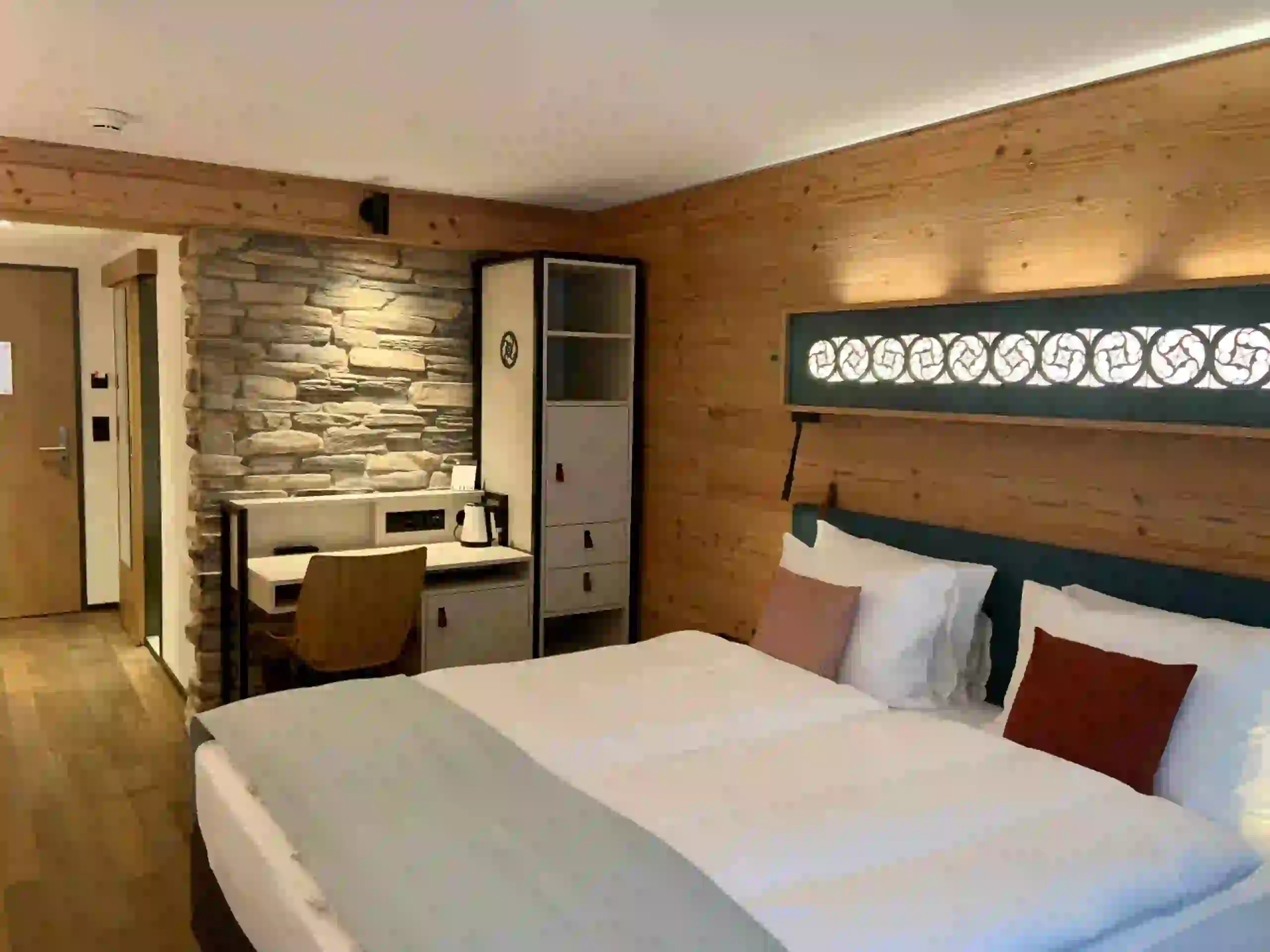 A cozy hotel room with a large bed, white linens, and brown accent pillows. There is a stone wall, a desk with a chair, shelves, and wood-paneled walls, giving the space a warm, inviting feel.