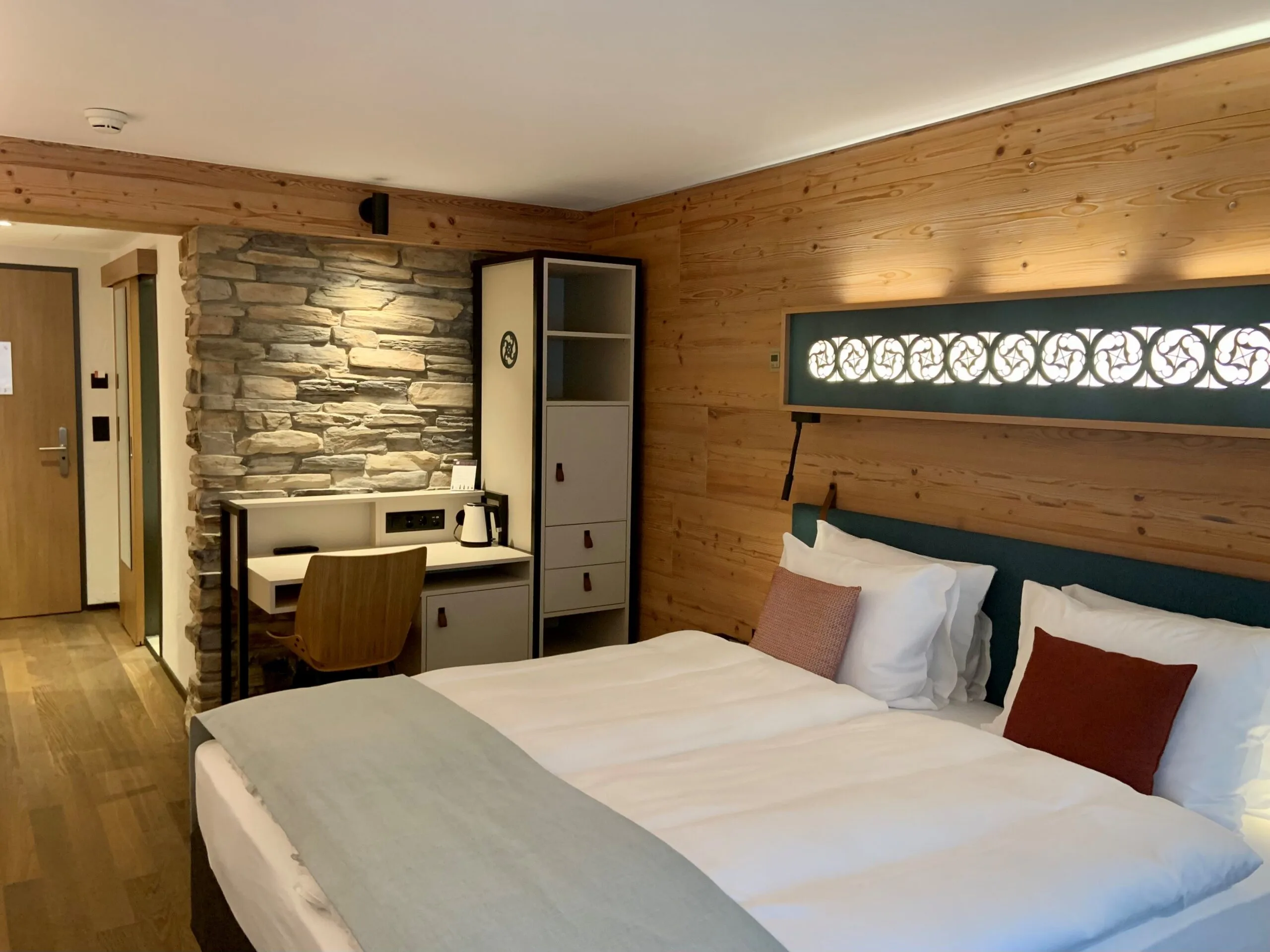 Ein gemütliches Hotelzimmer mit Doppelbett, weißer Bettwäsche und braunen Akzentkissen. Ein Holzschreibtisch mit Stuhl, ein Schrank und eine Steinwand verleihen dem Raum Charme. Warme Beleuchtung schafft eine einladende Atmosphäre.