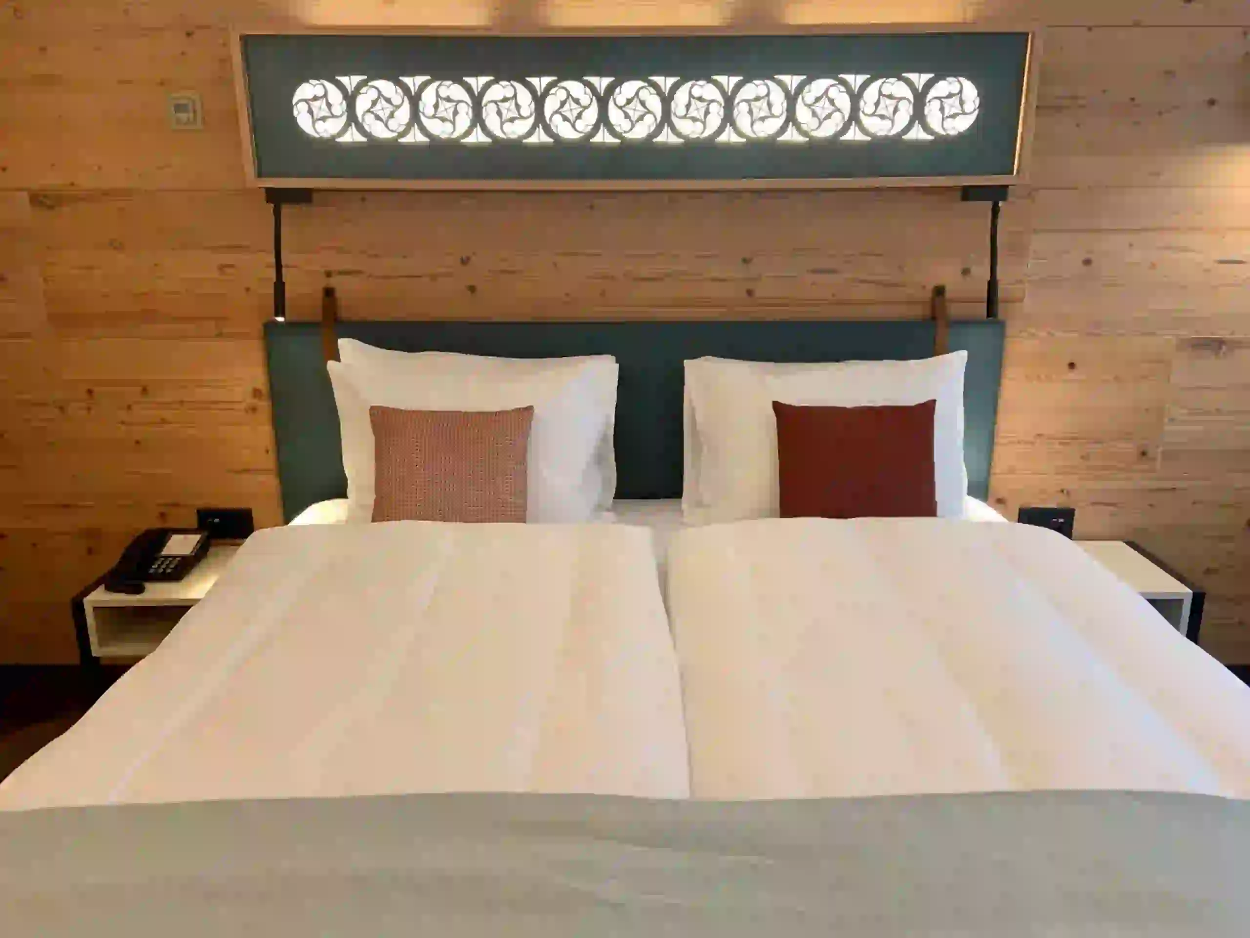 A neatly made hotel bed with two white duvets, two large pillows, and two decorative cushions. Wooden walls and a decorative panel with a swirl pattern are visible behind the bed. A phone sits on a bedside table.