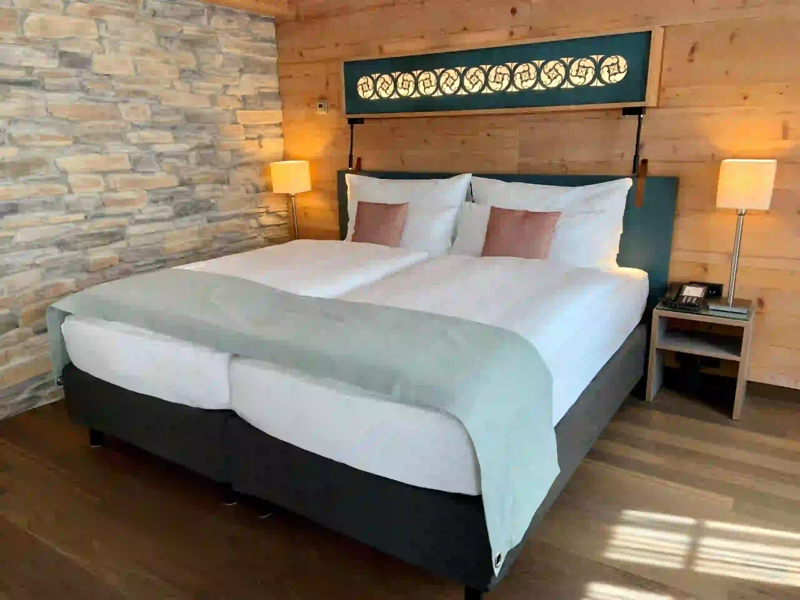 A cozy, modern bedroom with a large double bed, white linens, two pink accent pillows, and a light blue throw. The room features wood-paneled walls, a stone accent wall, bedside tables with lamps, and a decorative headboard.