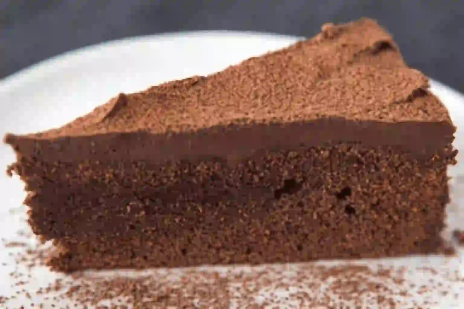Gros plan sur une tranche de gâteau au chocolat sur une assiette blanche, recouverte d'une couche de glaçage au chocolat et saupoudrée de poudre de cacao.