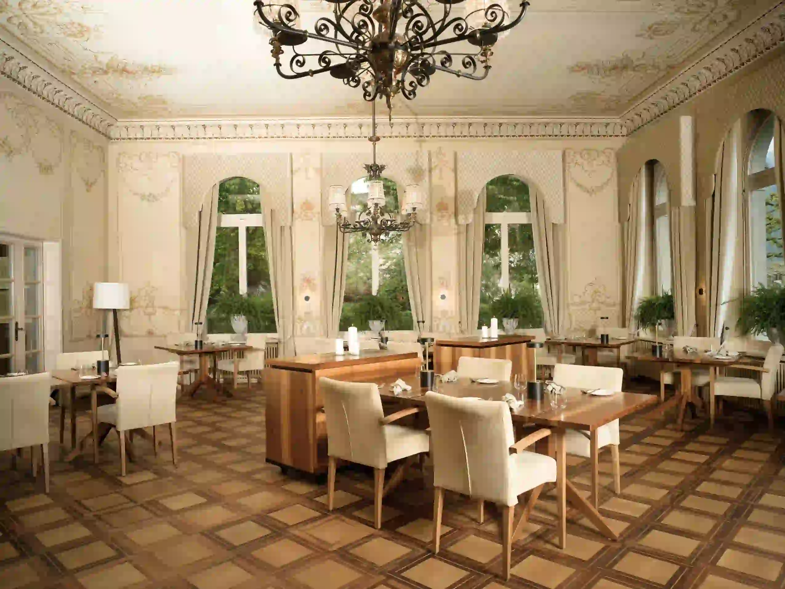 A dining room with high ceilings, decorative wall moldings, and chandeliers, featuring wooden tables and upholstered chairs arranged throughout the space.